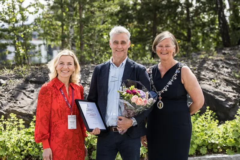 Prisvinner står i midten med blomster og innrammet pris fra Legeforeningen, sammen med to andre personer utendørs