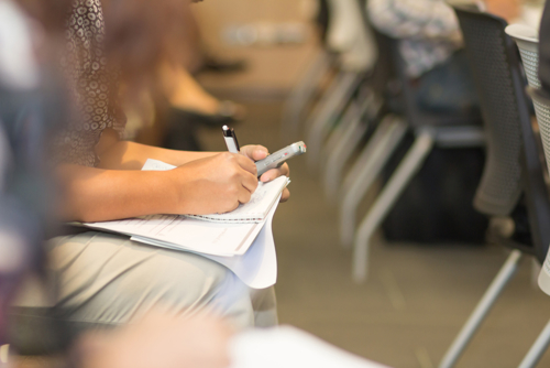 Kvinne som sitter på en stol i et klasserom og noterer på en notatblokk med penn.
